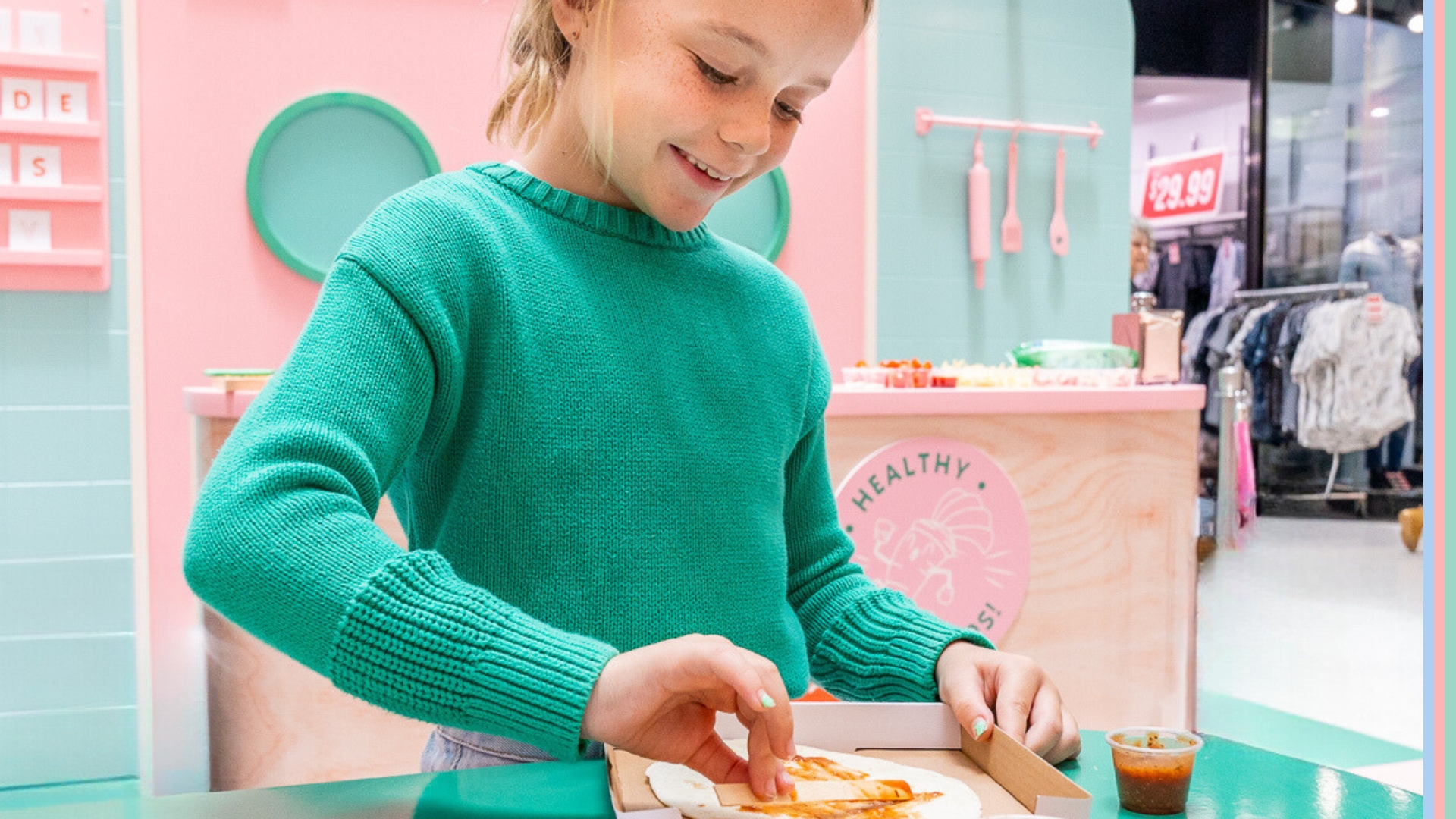 Girl dressed in green sweater smiling and holding a pizza box with a kitchen setup in the background